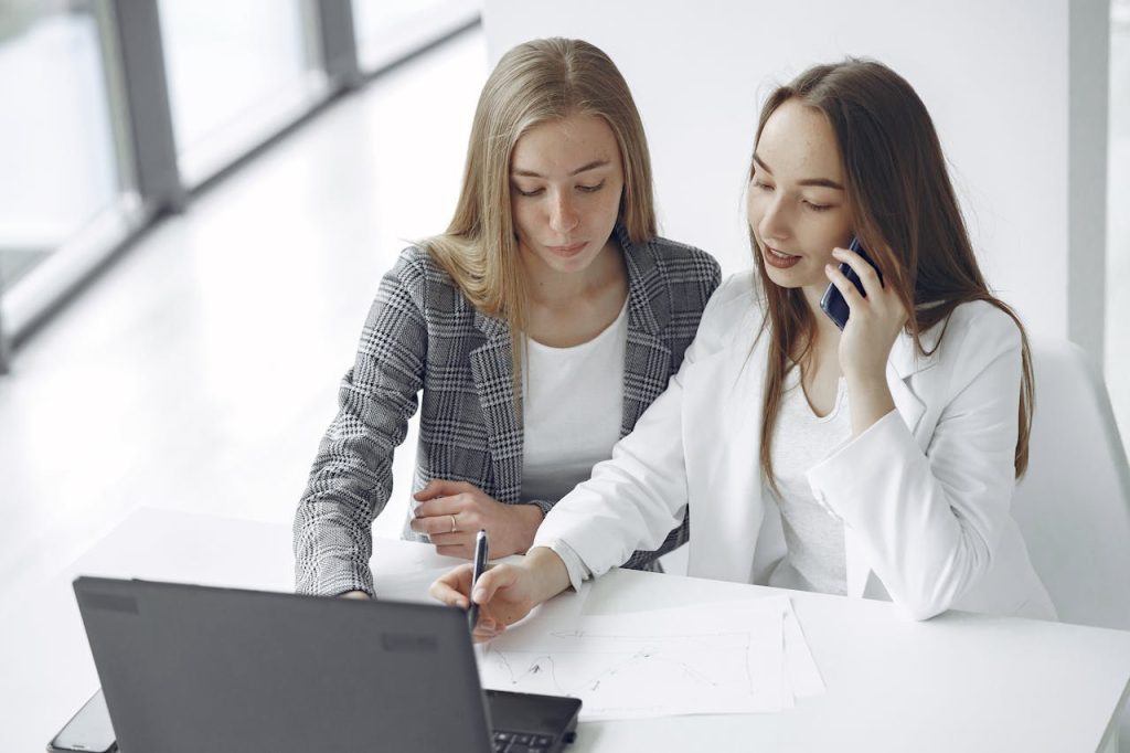 Two business women sitting on a desk working on a laptop 